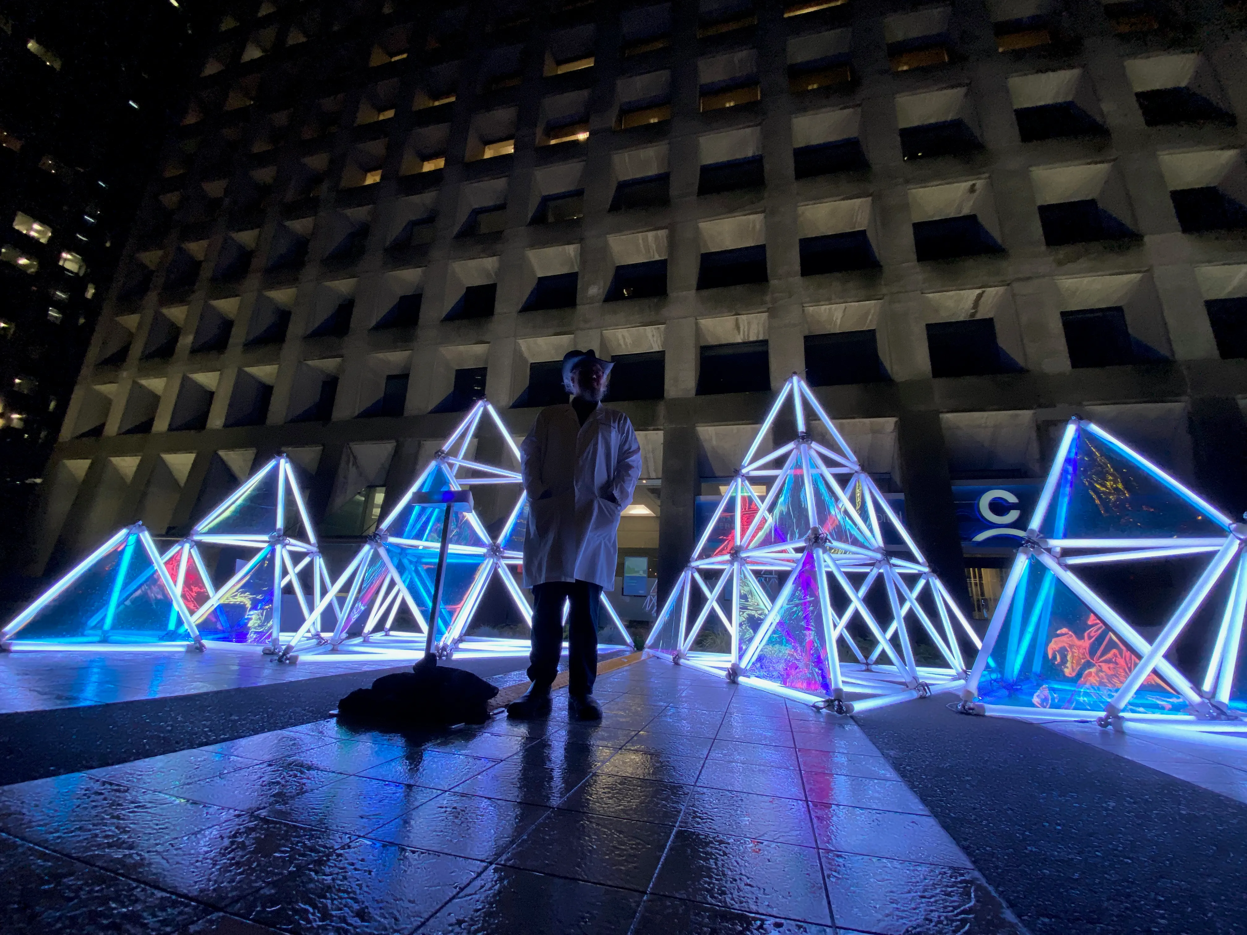 A person in a lab coat and cowboy hat stands at night on a rain-slicked plaza, silhouetted against glowing neon geometric sculptures in front of a stark, grid-like concrete building.